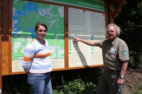 Henning Walter, Leiter des Nationalparkforstamtes Eifel, und Chantal van Marwijk, Leiterin des Fachgebiets Hoheit, zeigen eine der neuen Begrüßungstafeln an den Nationalparkzugängen.